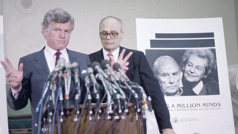 Dingell, standing behind former senator Ted Kennedy at an Alzehimer’s bill announcement, 1987.